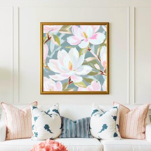 K&ouml;nnte beinhalten: Gerahmtes Blumenbild mit gro&szlig;en wei&szlig;en und rosa Magnolienbl&uuml;ten vor einem ged&auml;mpften blaugr&uuml;nen Hintergrund. Der goldene Rahmen erg&auml;nzt das Kunstwerk, das &uuml;ber einem wei&szlig;en Sofa mit dekorativen Kissen h&auml;ngt.
