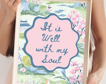 May include: A rectangular framed print featuring a watercolor-style landscape with green hills, pink cherry blossoms, and a blue building. In the center, a pink scalloped shape contains the dark blue text: It is Well with my Soul.