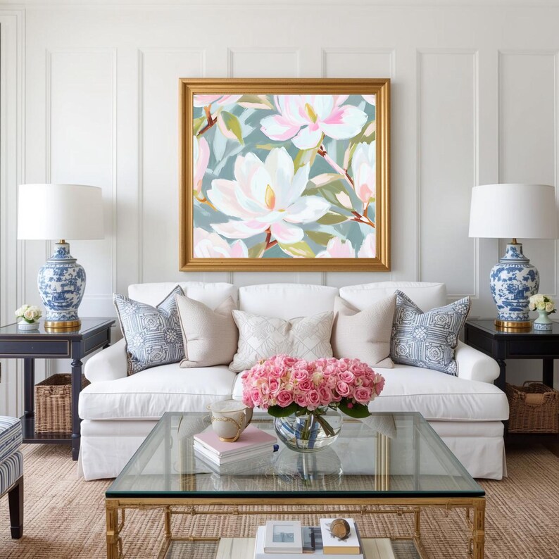 K&ouml;nnte beinhalten: Wohnzimmer mit wei&szlig;em Sofa, dekorativen Kissen und einem goldgerahmten Blumenbild. Ein Glastisch h&auml;lt eine Vase mit rosa Rosen. Blaue und wei&szlig;e Lampen flankieren das Sofa. Der Raum hat eine neutrale Farbpalette.