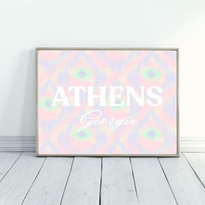 May include: A framed print with the word "ATHENS" in white block letters above "Georgia" in cursive. The background has a pastel pattern in pink, yellow, green, and blue. The frame is a light brown colour.