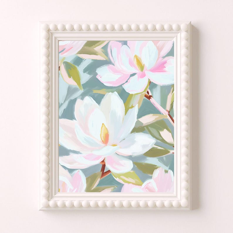 K&ouml;nnte beinhalten: Ein gerahmter Kunstdruck mit einem Aquarellgem&auml;lde von wei&szlig;en und rosa Magnolienbl&uuml;ten mit gelben Zentren und gr&uuml;nen Bl&auml;ttern vor einem tealfarbenen Hintergrund. Der wei&szlig;e Rahmen hat einen Perlenrand.