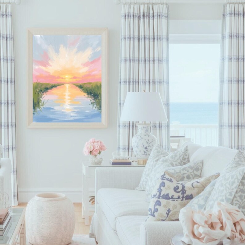 May include: A framed landscape painting depicts a sunset over a waterway, with pink, yellow, and blue hues. The artwork hangs above a white sofa with patterned pillows. The room features white walls, curtains, and a decorative vase.