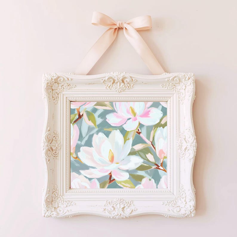 K&ouml;nnte beinhalten: Ein gerahmter Blumendruck mit wei&szlig;en und rosa Magnolienbl&uuml;ten vor einem teal-farbenen Hintergrund. Der wei&szlig;e, verzierte Rahmen wird von einem hellrosa Band gehalten, das zu einer Schleife gebunden ist, was eine zarte und elegante &Auml;sthetik erzeugt.