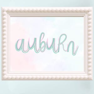 May include: A white framed print with the word "auburn" in a layered script font. The word is in a teal and pink color scheme, set against a soft watercolor background. The frame has a scalloped edge.