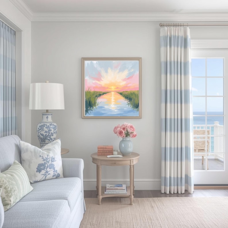 May include: A living room scene featuring a framed landscape painting with a sunset over water. A light blue sofa with floral pillows, a round wooden side table with a vase of pink roses, and striped curtains complete the coastal decor.