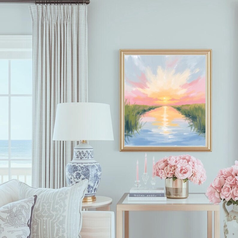 May include: A framed landscape painting depicts a sunset over a waterway, with pink, yellow, and blue hues. The painting is in a gold frame, hanging above a table with a floral arrangement and candles. A blue and white patterned lamp sits nearby.