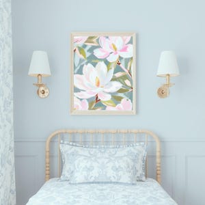 K&ouml;nnte beinhalten: Ein gerahmter Kunstdruck mit gro&szlig;en, wei&szlig;en und rosa Magnolienbl&uuml;ten vor einem ged&auml;mpften t&uuml;rkisblauen Hintergrund. Das Kunstwerk wird &uuml;ber einem Bett mit einem hellen Kopfteil und passender Bettw&auml;sche ausgestellt. Zwei goldene Wandleuchten mit wei&szlig;en Schirmen flankieren das Kunstwerk.