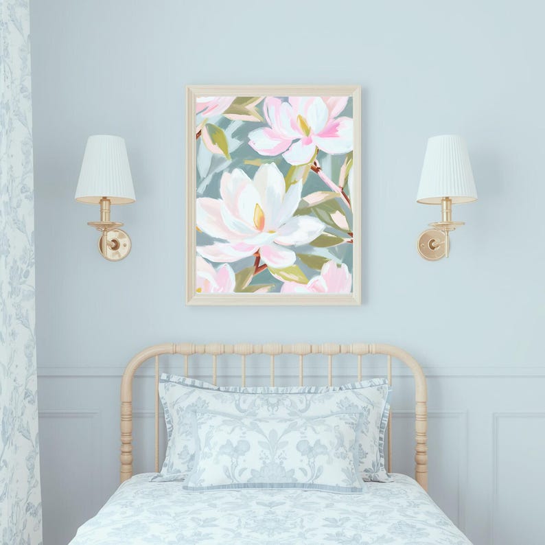K&ouml;nnte beinhalten: Ein gerahmter Kunstdruck mit gro&szlig;en, wei&szlig;en und rosa Magnolienbl&uuml;ten vor einem ged&auml;mpften t&uuml;rkisblauen Hintergrund. Das Kunstwerk wird &uuml;ber einem Bett mit einem hellen Kopfteil und passender Bettw&auml;sche ausgestellt. Zwei goldene Wandleuchten mit wei&szlig;en Schirmen flankieren das Kunstwerk.