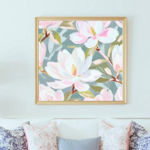 K&ouml;nnte beinhalten: Gerahmtes Blumenbild mit gro&szlig;en wei&szlig;en und rosa Magnolienbl&uuml;ten vor einem ged&auml;mpften blau-gr&uuml;nen Hintergrund. Der goldfarbene Rahmen betont das Kunstwerk, das &uuml;ber einem wei&szlig;en Sofa mit Zierkissen h&auml;ngt.