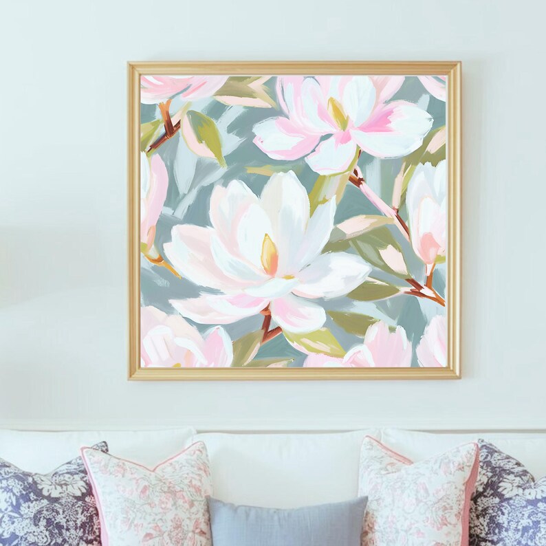 K&ouml;nnte beinhalten: Gerahmtes Blumenbild mit gro&szlig;en wei&szlig;en und rosa Magnolienbl&uuml;ten vor einem ged&auml;mpften blau-gr&uuml;nen Hintergrund. Der goldfarbene Rahmen betont das Kunstwerk, das &uuml;ber einem wei&szlig;en Sofa mit Zierkissen h&auml;ngt.