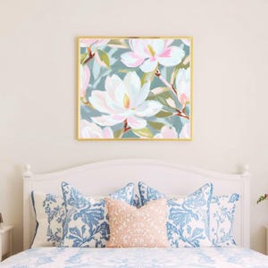 K&ouml;nnte beinhalten: Gerahmter Blumendruck mit wei&szlig;en und rosa Magnolienbl&uuml;ten auf einem blaugr&uuml;nen Hintergrund. Das Kunstwerk befindet sich in einem goldenen Rahmen, &uuml;ber einem Bett mit blau und pfirsichfarben gemusterten Kissen. Dekorativer Stil.
