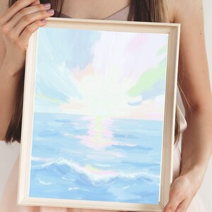 May include: A framed art print depicting a seascape with a sunset. The artwork features a light blue ocean with white waves, a bright yellow sun, and a sky with pastel colors. The frame is a light beige color.