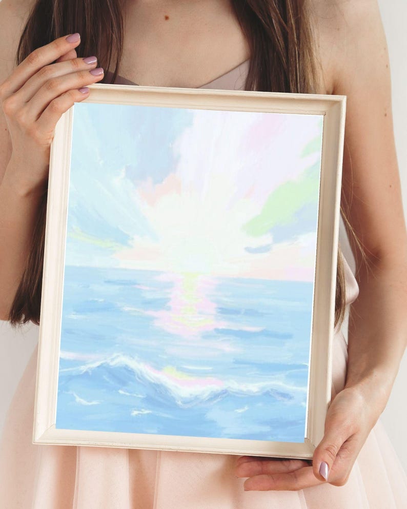 May include: A framed art print depicting a seascape with a sunset. The artwork features a light blue ocean with white waves, a bright yellow sun, and a sky with pastel colors. The frame is a light beige color.