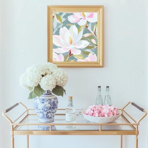 K&ouml;nnte beinhalten: Ein gerahmtes Blumenbild mit goldenem Rahmen h&auml;ngt &uuml;ber einem goldenen Servierwagen. Der Wagen enth&auml;lt eine blau-wei&szlig;e Vase mit wei&szlig;en Blumen, eine Flasche, eine Schale mit rosa Bl&uuml;tenbl&auml;ttern und zwei Wasserflaschen. Das Gem&auml;lde zeigt eine gro&szlig;e wei&szlig;e Blume.