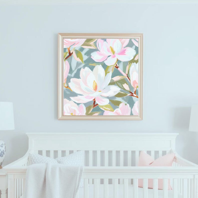 K&ouml;nnte beinhalten: Ein gerahmter Kunstdruck mit einem floralen Muster aus gro&szlig;en, wei&szlig;en und rosa Magnolienbl&uuml;ten mit gelben Zentren und gr&uuml;nen Bl&auml;ttern auf einem ged&auml;mpften tealfarbenen Hintergrund. Das Kunstwerk h&auml;ngt &uuml;ber einem wei&szlig;en Kinderbett.
