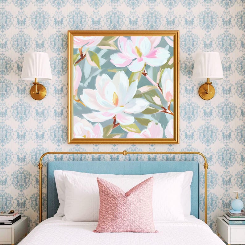 K&ouml;nnte beinhalten: Ein gerahmter Kunstdruck mit wei&szlig;en und rosa Magnolienbl&uuml;ten auf t&uuml;rkisblauem Hintergrund. Der goldene Rahmen h&auml;ngt &uuml;ber einem Bett mit blauem Kopfteil, wei&szlig;er Bettw&auml;sche und einem rosa Kissen. Zwei goldene Wandleuchten flankieren das Kunstwerk.