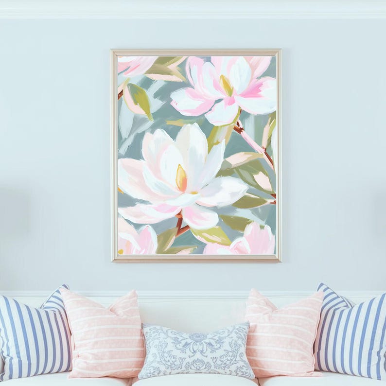 K&ouml;nnte beinhalten: Gerahmtes Blumenbild mit gro&szlig;en, stilisierten Magnolienbl&uuml;ten in Wei&szlig;- und Rosat&ouml;nen vor einem ged&auml;mpften t&uuml;rkisblauen Hintergrund. Das Kunstwerk befindet sich in einem goldenen Rahmen und h&auml;ngt &uuml;ber einem wei&szlig;en Sofa mit dekorativen Kissen.