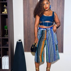 May include: A woman wearing a blue denim crop top with cutouts and a striped, multi-colored, high-waisted culottes. She is wearing gold sandals and a black handbag with a gold chain.