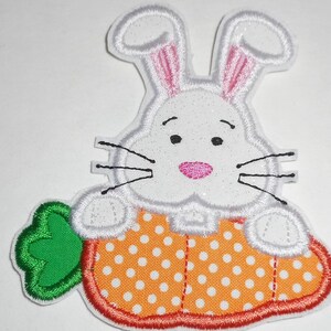May include: White bunny rabbit with pink ears holding a carrot with orange and white polka dots. The carrot has a green leaf.