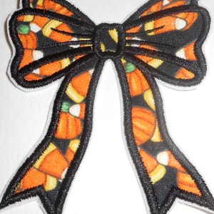 May include: A Halloween-themed bow applique with a black outline and a candy corn pattern. The bow features orange, yellow, and white candy corn on a black background, perfect for festive crafts and decorations.