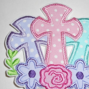 May include: An embroidered patch with three crosses in pink, purple, and blue with white polka dots. The crosses are surrounded by green leaves and pink, purple, and blue flowers.