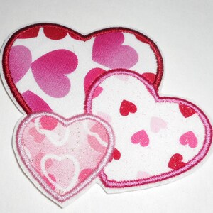 May include: Three pink and red felt hearts with white backgrounds and different patterns of smaller hearts. The hearts are layered on top of each other.