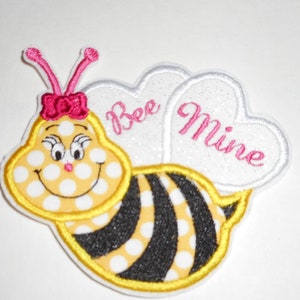 May include: A yellow and black bee with white polka dots and a pink bow. The bee has a smiling face and is holding two white hearts that say "Bee Mine" in pink lettering.