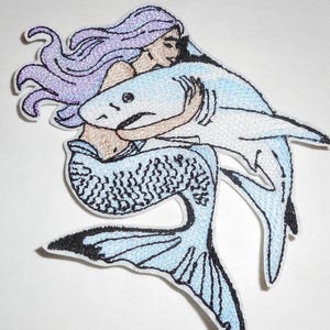 May include: An embroidered patch featuring a figure with long lavender hair and a mermaid tail embracing a light blue shark. The design uses black outlines to define the shapes of the mermaid and the shark, with a white border surrounding the entire image.