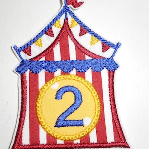 May include: Embroidered patch featuring a circus tent design with red and white stripes, blue trim, and a yellow circle with the number 2. The patch has a red flag at the top and small triangular flags along the top edge.