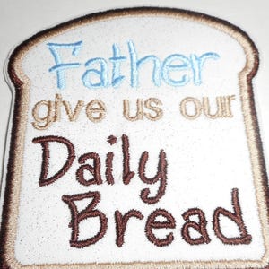 May include: Embroidered patch in the shape of a slice of bread. The patch features the words "Father give us our Daily Bread" in blue and brown thread, with a brown border.