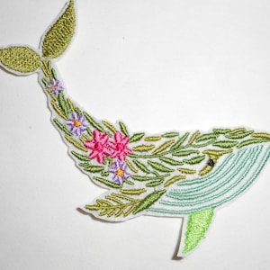 May include: Embroidered patch featuring a whale design. The whale is decorated with green leaves, pink and purple flowers, and green and blue stitching. The patch has a white border and is set against a white background.
