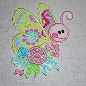 May include: A pink and green embroidered butterfly with a floral design. The butterfly has a pink face with black eyes and a pink smile. The flowers are pink, blue, and green.