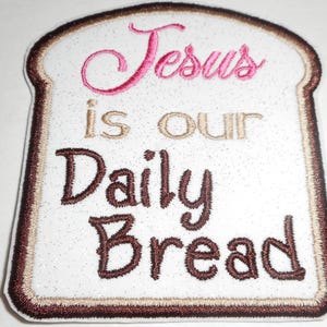 May include: Embroidered patch in the shape of a slice of bread. The patch features the words "Jesus is our Daily Bread" in pink, tan, and brown thread. The bread slice has a brown crust and a white center.