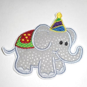 May include: A gray embroidered elephant wearing a colorful party hat. The elephant has a red, green, and yellow patterned saddle.