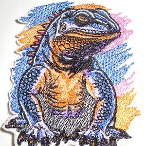 May include: An embroidered patch featuring a detailed lizard with textured scales in shades of blue, orange, and black. The lizard is depicted in a seated position against a background of scribbled, multi-colored lines in blue, orange, and pink.