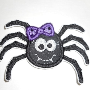 May include: Embroidered patch featuring a cartoon spider with a black body, eight legs, and a purple bow. The spider has large, white eyes and a smiling mouth. The patch has a white border.