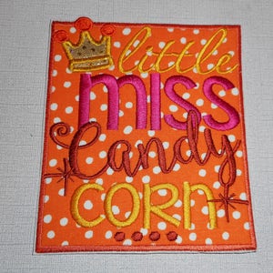 May include: Orange and white fabric with white polka dots. The fabric has an embroidered design that says "little miss candy corn" with a gold crown above the text.