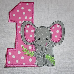 May include: An embroidered applique featuring the number one in pink fabric with white polka dots, outlined in dark pink thread. A gray elephant with white polka dots and pink inner ears is positioned in front, holding a green leafy branch.