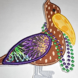 May include: Embroidered pelican patch with a brown body, white polka dots, and a yellow beak with orange polka dots. The patch features a purple and green wing, and green and purple bead strands.
