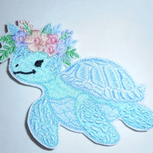 May include: An embroidered patch featuring a light blue sea turtle with a textured shell and a smiling face. The turtle wears a crown of colorful flowers in shades of pink, purple, and blue, accented with green leaves, all on a white background.