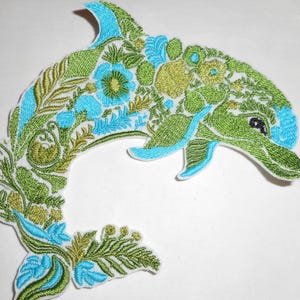May include: An embroidered iron-on patch shaped like a leaping dolphin. The design features a detailed floral and botanical pattern in shades of lime green, olive green, and bright sky blue, with a small black eye visible on the dolphin head.