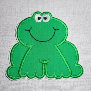 May include: Green felt applique of a smiling frog with black eyes and a white outline. The frog is sitting with its legs crossed.