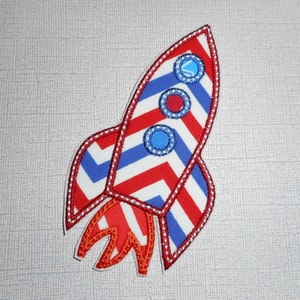 May include: Red, white, and blue rocket ship applique with a chevron pattern. The rocket has three round windows with blue and white trim.