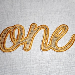 May include: A gold-colored embroidered patch with the word "one" in a cursive font. The word is outlined in a darker gold thread, with a lighter gold mesh and sequins filling the letters. The patch is on a light gray textured background.