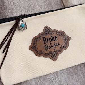 May include: A cream-coloured canvas zip pouch featuring a brown leather-look patch with the text Broke & Boujee in black. The black zip pull is adorned with a silver-toned charm containing a small turquoise stone and two brown leather cords.