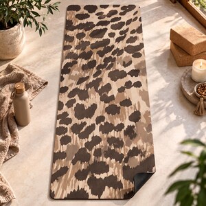 May include: A rectangular yoga mat featuring a repeating leopard print pattern in shades of dark brown, tan, and beige. The mat has rounded corners and a smooth surface texture, with the dark brown pattern layered over a lighter tan background.
