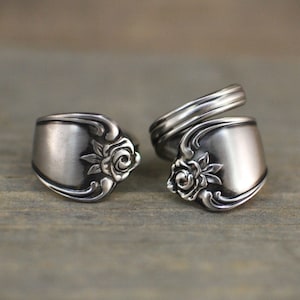 Op de afbeelding: Twee zilveren lepelringen met een bloemenrozenontwerp. De ringen zijn gemaakt van hergebruikt zilverwerk en hebben een vintage, handgemaakte uitstraling. De ringen worden gepresenteerd op een houten oppervlak.