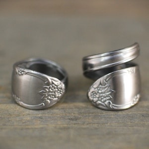 Op de afbeelding: Twee zilveren ringen gemaakt van hergebruikt bestek. Beide ringen zijn breed en hebben een bloemmotief. De ring aan de linkerkant is een enkele band, terwijl de ring aan de rechterkant een dubbele band is.