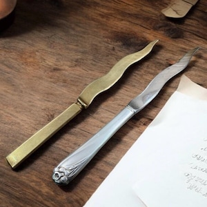 Op de afbeelding: Twee briefopeners liggen op een houten oppervlak. Eén is messingkleurig met een rechte handgreep en een gebogen lemmet. De andere is zilverkleurig met een bloemmotief op de handgreep en een gebogen lemmet. Een handgeschreven notitie is gedeeltelijk zichtbaar.