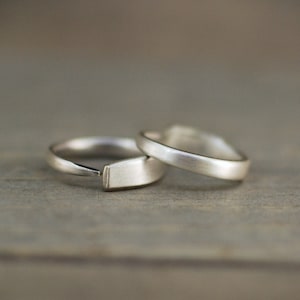 Op de afbeelding: Twee zilveren ringen, een met een platte, rechthoekige band en de andere met een ronde band, liggen op elkaar gestapeld op een houten oppervlak.